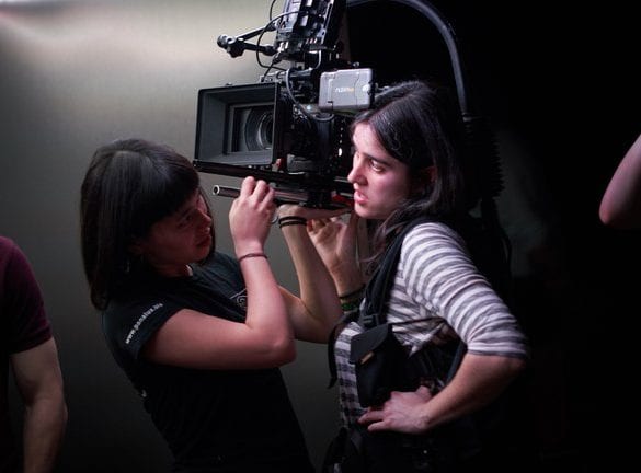 A woman with medium length black hair stands wearing a camera rig with a large digital camera on her right shoulder. An Assistand Camera woman with a navy t-shirt and bobbed brown hair is asisting her by wiping the camera lense.