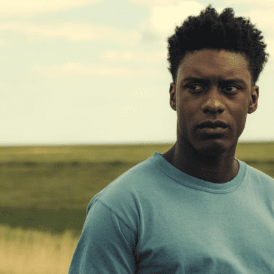 A young Black man with a light blue t-shirt stands in a field looking pensive.