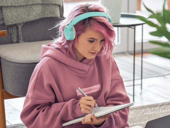 A white woman with pink hair, light blue headphones and a pink jumper is sat watching something on a laptop taking notes in a notepad