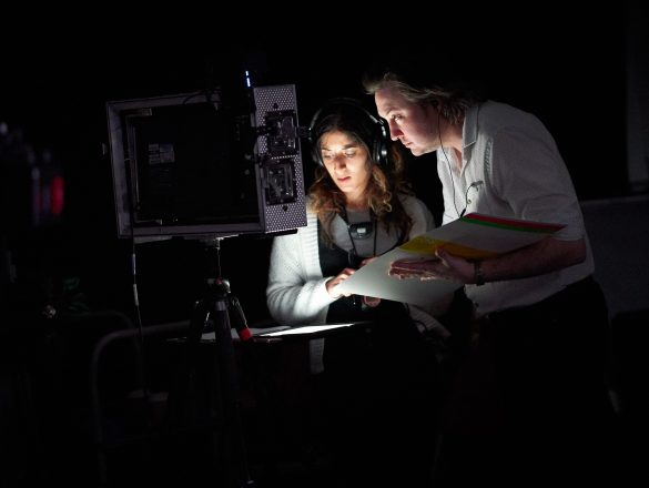 A woman and a man stand in front of a minitor which illuminates their faces. They are both holding scripts.