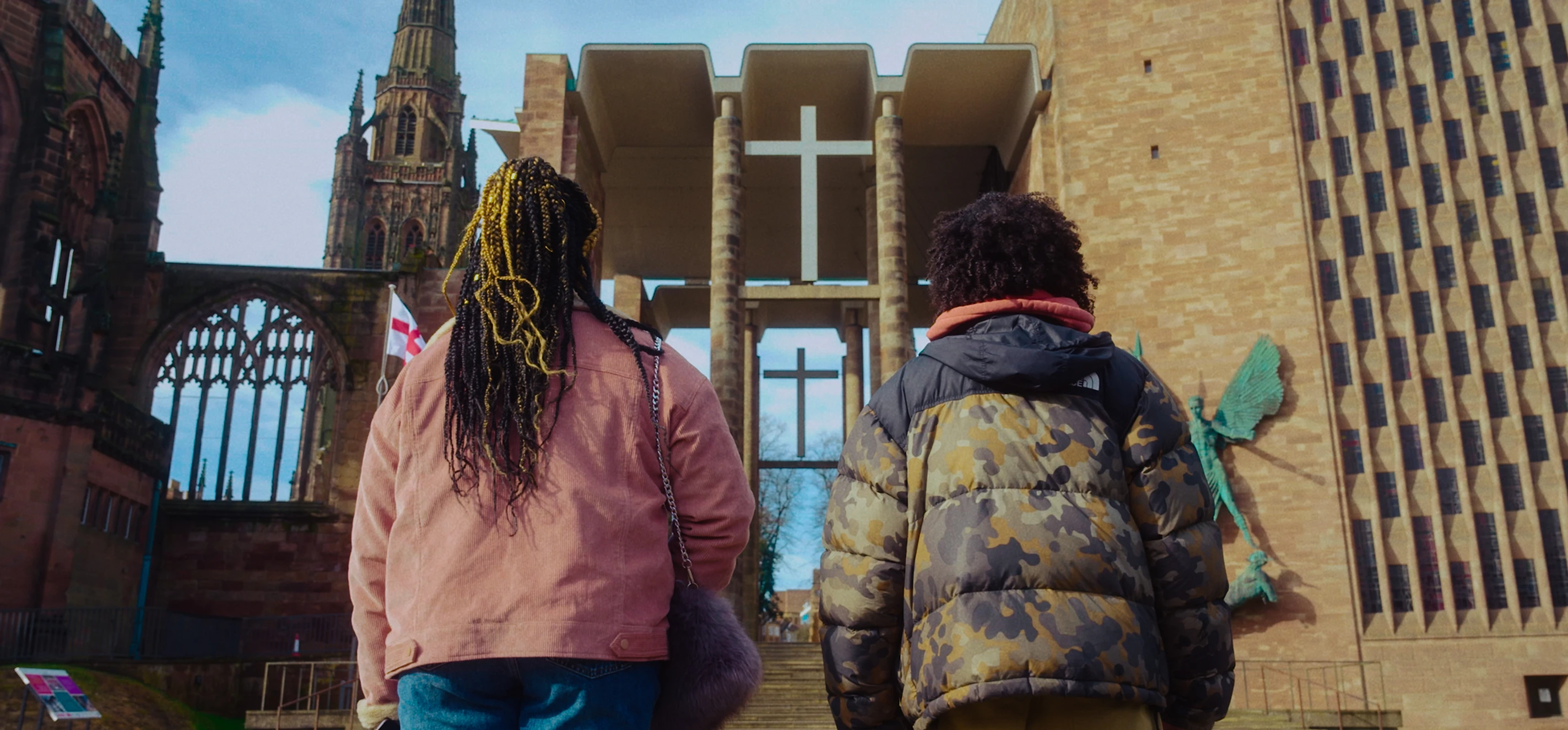 Two young people stand next to each other looking up at Coventry Cathedral. One has a denim jacket and black and blond braids half down and half in a bun and the other has a khaki puffer jacked and natural dark brown curls.