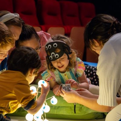 A young girl surrounded by fairy lights and wearing a googly eye mask with parents