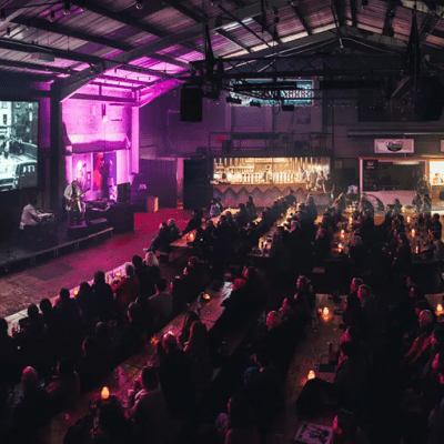 A large crowd sits in an auditorium watching a screen, which is showing a black and white image of traffic. Two musicians are in front of the screen, both dressed in white lab coats.