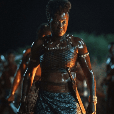 Viola Davis stands square-shouldered and looking prepared to fight in front of a larger group of black women (out of focus in the background) - she holds a weapon and wears a decorative necklace..