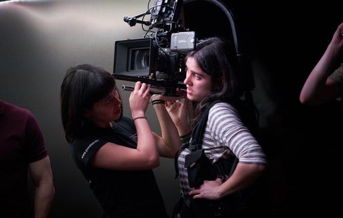 A woman with medium length black hair stands wearing a camera rig with a large digital camera on her right shoulder. An Assistand Camera woman with a navy t-shirt and bobbed brown hair is asisting her by wiping the camera lense.