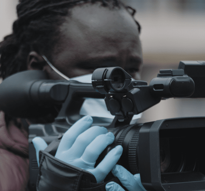 A Black man with a medical face mask is holding a camera with gloes and looking down the view finder
