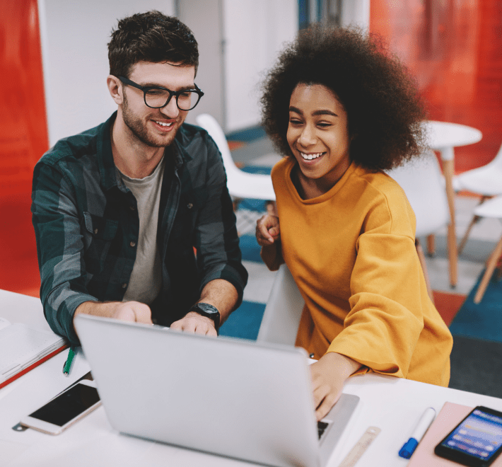 A Black woman with natural brown hair and wearing a yellow top and a white man with short brown hair, glasses and a green checked shirt sit in front of a laptop in an office with red walls and white furniture.