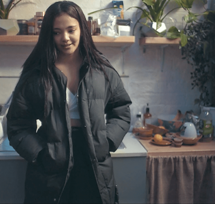 A young woman stands in a kitchen, wearing a long black jacket with her hands in her pockets, and smiling slightly.