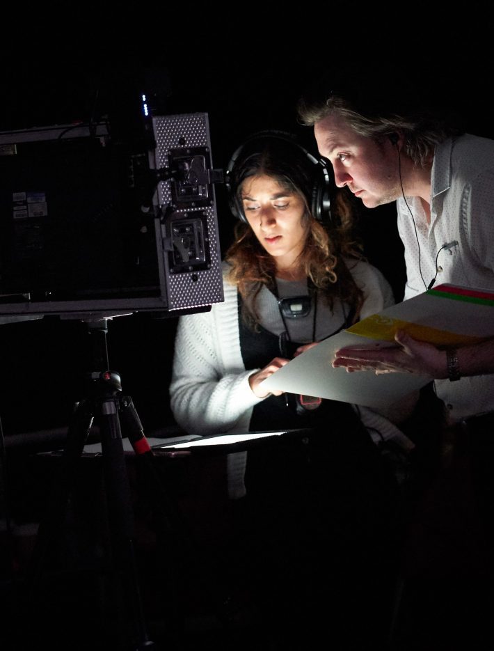 A woman and a man stand in front of a minitor which illuminates their faces. They are both holding scripts.