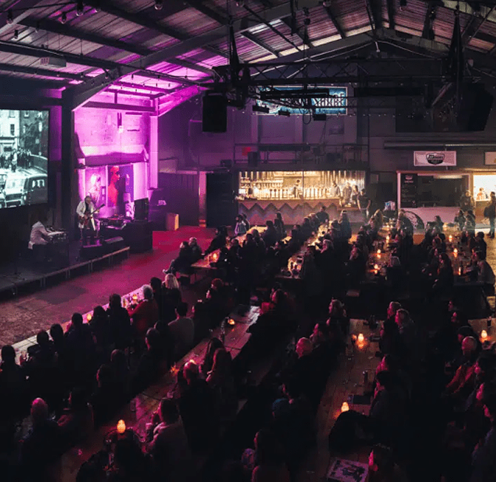 A large crowd sits in an auditorium watching a screen, which is showing a black and white image of traffic. Two musicians are in front of the screen, both dressed in white lab coats.