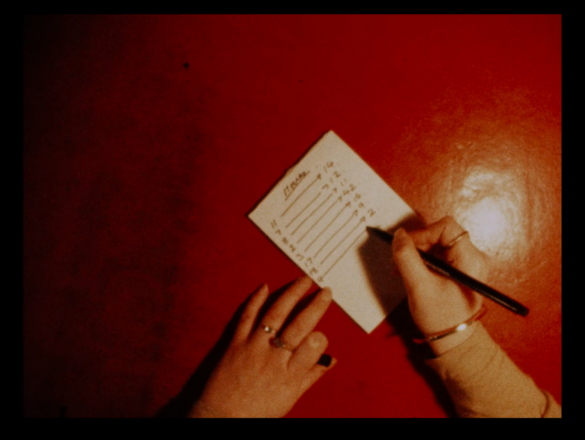 Hands coming from the bottom of the image write on a peice of paper placed on a red table