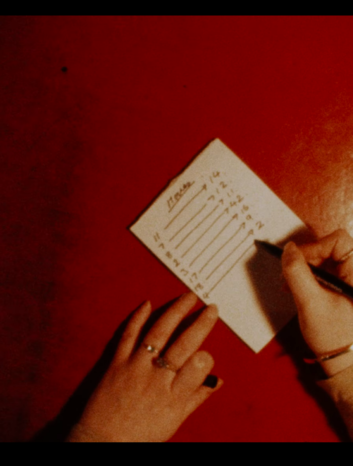 Hands coming from the bottom of the image write on a peice of paper placed on a red table