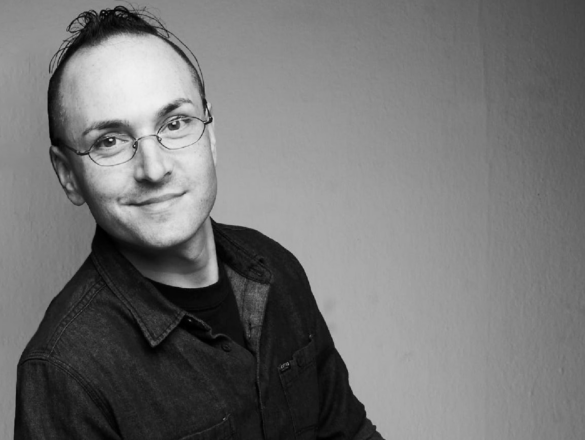 A man in a black Tshirt wearing glasses and looking in the camera lens leans against a white wall