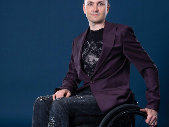 Owen dressed in a purple blazer and trousers sits in their wheelchair looking towards the camera with a blue backdrop