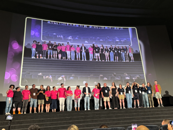 A large group of industry personnel at Deaffest stand on stage some are wearing bright pink tshirts and all looking around in different directions candid