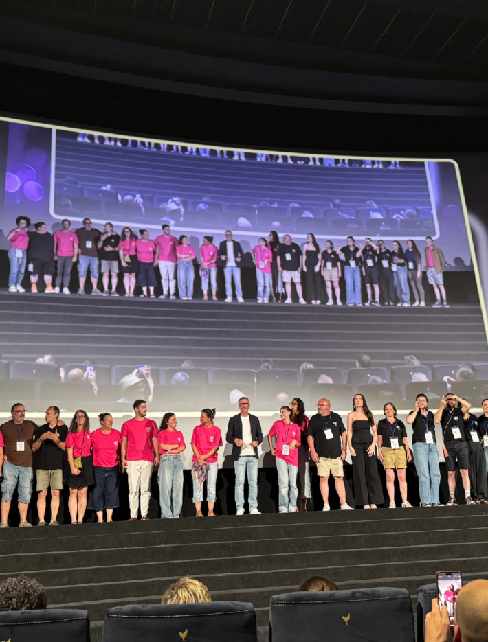 A large group of industry personnel at Deaffest stand on stage some are wearing bright pink tshirts and all looking around in different directions candid
